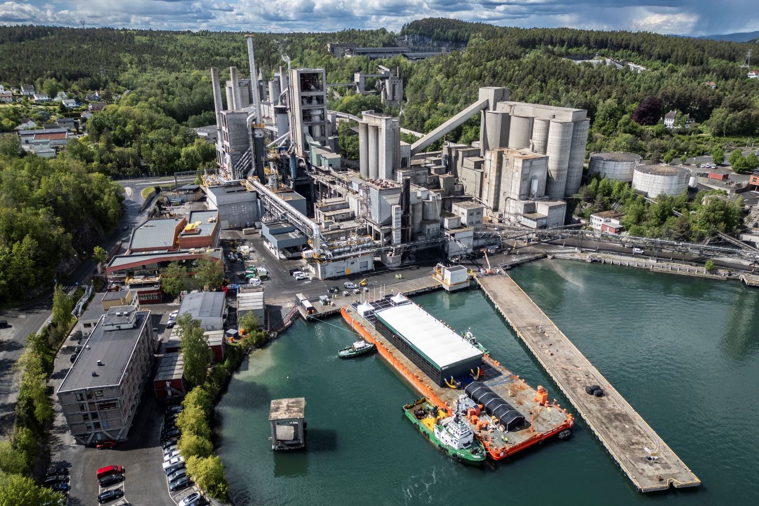 Aerial view of a cement plant with silos, conveyors, and processing units, located near a port with a docked barge for material transport.
