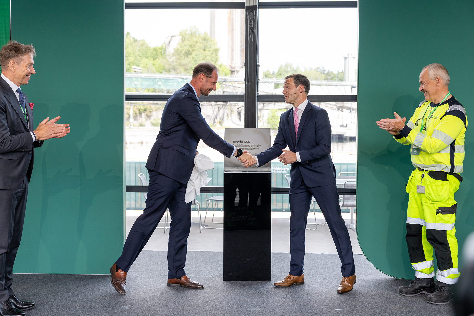 Crown Prince Haakon and Dr Dominik von Achten in suits shake hands at the center of a stage in front of a plaque, with the Crown Prince holding a white cloth. On the left, a man in a suit applauds, and on the right, a worker in high-visibility gear claps. Large windows in the background reveal a seaside view with trees and buildings.