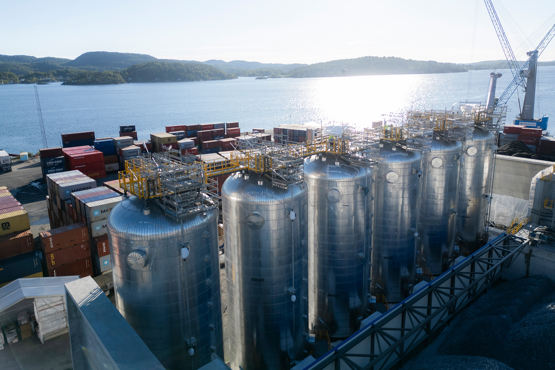 Six large, silver industrial storage tanks aligned in a row at a port, surrounded by shipping containers and cranes, with a body of water and distant hills in the background under bright sunlight.
