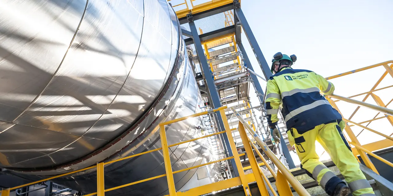 A worker in high-visibility safety gear and a helmet ascends a yellow metal staircase next to large cylindrical industrial equipment under clear blue sky.