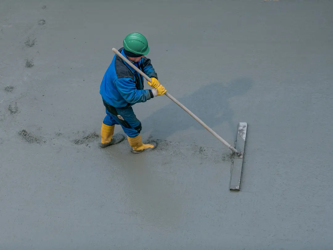 Worker in safety gear levelling wet concrete with a long-handled tool on a construction site.