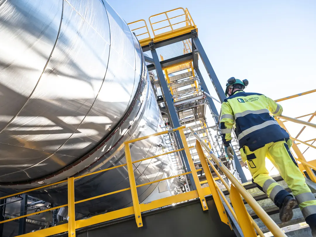 Muncitor în echipament de protecție galben urcă pe o scară galbenă spre o platformă industrială, lângă un rezervor metalic mare, circular, într-un mediu industrial iluminat de soare.